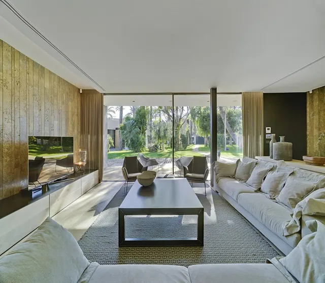 Salón con paneles de madera y gran ventanal al jardín de palmeras